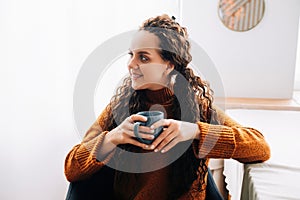 Young smiling pretty woman holding cup drinking warm tea or coffee relaxing dreaming at home. Happy positive calm lady enjoying