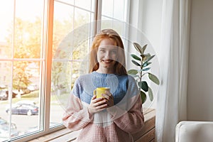 Young smiling red hair woman in comfy knitted warm sweater holding yellow coffee cup