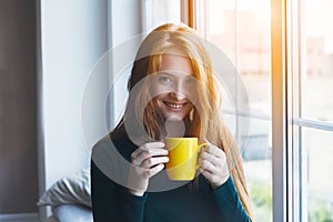 Young smiling red hair woman holding yellow coffee cup, enjoying morning coffee