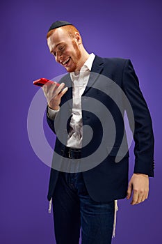 Young smiling redhead emotional Jewish man looking on phone, greeting with holiday against purple studio background