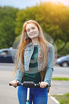 Young smiling redhead female student riding blue electric kick scooter