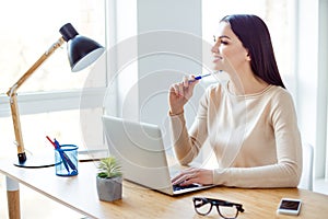 Young smiling successful businesswoman sitting at the table with laptop and think about new project