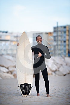 Young smiling surfer in black zipless surfing wetsuit with shortboard showing shaka sign in Costa Da Caparica, Portugal photo