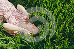 Young smiling tender beautiful woman with closed eyes in light patterned dress lying on grass resting in sunny weather