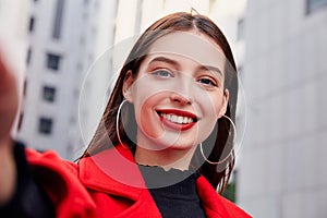 Young, smiling and unsurpassed girl with large round earrings takes a selfie against a gray building