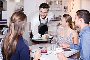 young smiling waiter bringing ordered dishes to friends in tearoom