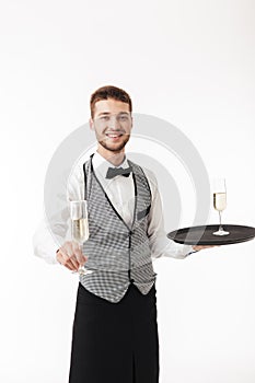 Young smiling waiter in uniform joyfully holding tray while offering glass of champagne on camera over white background