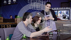 Young smiling waitress standing in cafe near the table of happy couple and putting their order into digital tablet. Man