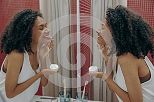 Young smiling woman applying moisturising face cream near mirror In bathroom. Skin care concept