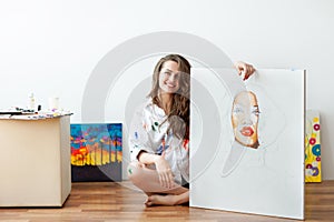 Young smiling woman artist sits on floor with unfinished masterpiece