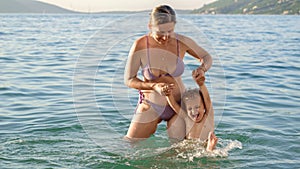 Young smiling woman in bikini holding her baby son by hands while having fun in the sea. Summertime, holiday, vacation