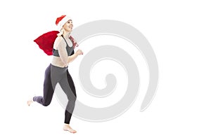 Young smiling woman in a black tracksuit in a Santa Claus hat and a red bag with gifts runs. Full height. Isolated over white