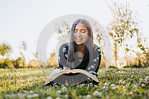 Young smiling woman with braces, believer, studying her bible while lying on the grass, in a natural space.