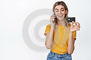 Young smiling woman calling someone, talking on mobile phone and showing credit card, order something, standing over