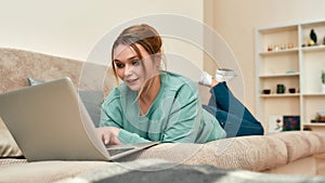 Young smiling woman in casual clothes lying on the sofa in the living room and working on laptop