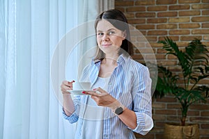 Young smiling woman with cup of morning coffee at home near window