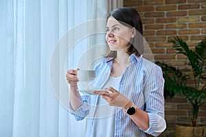 Young smiling woman with cup of morning coffee at home near window