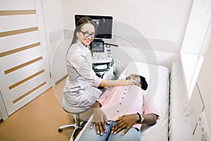 Young smiling woman doctor in diagnostic center making ultrasound thyroid gland test to female African patient. Woman