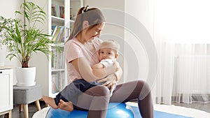 Young smiling woman doing fitness at home on fit ball while holding her little baby son