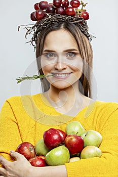 Young smiling woman with fruit. A beautiful brunette in a yellow sweater with purple grapes on her head, red and green apples in