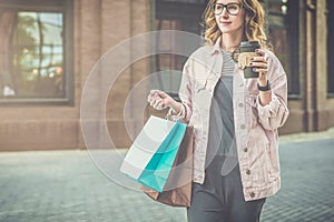 Young smiling woman in glasses carries shopping bags and holds cup of coffee.Girl bought presents for her friend.