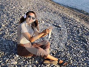 Young smiling woman in headphones and dark glasses sits on pebble coast and looks at the viewer