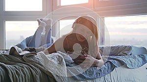 Young smiling woman in headphones relaxing with her lovely Maine Coon cat listens to music laying in bed by the window