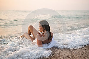 Young smiling woman holding glass of white wine lying in sea on sand coastline outdoor at sunset.