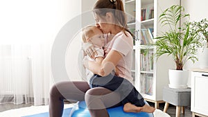 Young smiling woman holding her upset baby son and doing exercises on fitball at home. Concept of healthcare, sports and yoga at