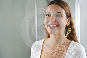 Young smiling woman holds a bamboo toothbrush looking herself in the mirror at home. Dental care, eco-friendly bamboo toothbrush,