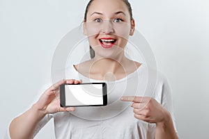 A young smiling woman holds a smartphone horizontally with a white screen with a place for copyspace and points at him the finger