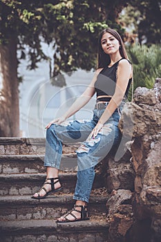 Young smiling woman jeans and accessories. Outdoors sitting on a low wall of rock on a staircase. Long hair