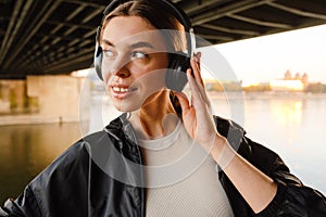 Young smiling woman listening music with headphones while standing on embankment