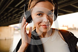 Young smiling woman listening music with headphones while standing on embankment