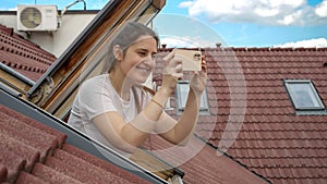 Young smiling woman looking out of the open attic window and making photos on her smartphone. Concept of tourism