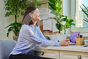 Young smiling woman looking out the window at home, sitting at desk