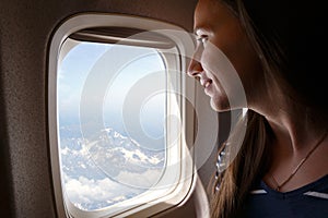 Young smiling woman looking through the plane window on the Alps mountains