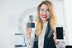 Young smiling woman manager stands in her bright office with bank card in one hand, showing smartphone in another hand ,