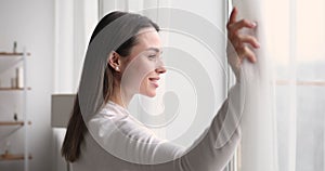 Young smiling woman opening curtain looking through window