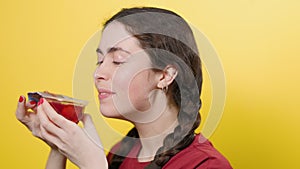 A young smiling woman opens a jar of jelly, sniffs it, and smells a terrible smell. Yellow background. The concept of junk food.