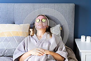 A young smiling woman with pink clay facial mask and cucumber slices making a refreshing eye mask in bedroom. Natural