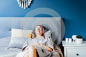 A young smiling woman with pink clay facial mask and cucumber slices making a refreshing eye mask in bedroom. Natural