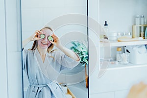 A young smiling woman with pink clay facial mask holds cucumber slices making a refreshing eye mask in bathroom. Natural