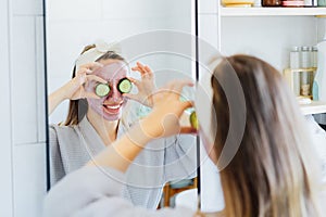 A young smiling woman with pink clay facial mask holds cucumber slices making a refreshing eye mask in bathroom. Natural