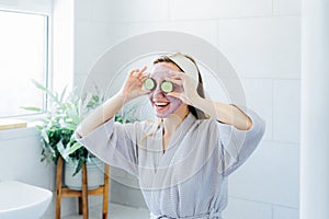 A young smiling woman with pink clay facial mask holds cucumber slices making a refreshing eye mask in bathroom. Natural