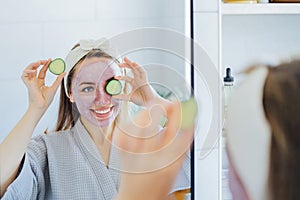 A young smiling woman with pink clay facial mask holds cucumber slices making a refreshing eye mask in bathroom. Natural