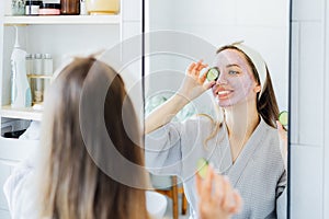 A young smiling woman with pink clay facial mask holds cucumber slices making a refreshing eye mask in bathroom. Natural