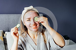 A young smiling woman with pink clay facial mask holds cucumber slices making a refreshing eye mask in bedroom. Natural