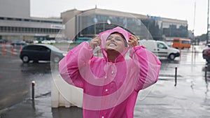 Young smiling woman with a pink raincoat while enjoying a walk through the city on a rainy day.