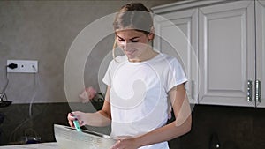 Young smiling woman prepares dough mixing ingredients in the the bowl using whisk in the kitchen. Homemade food